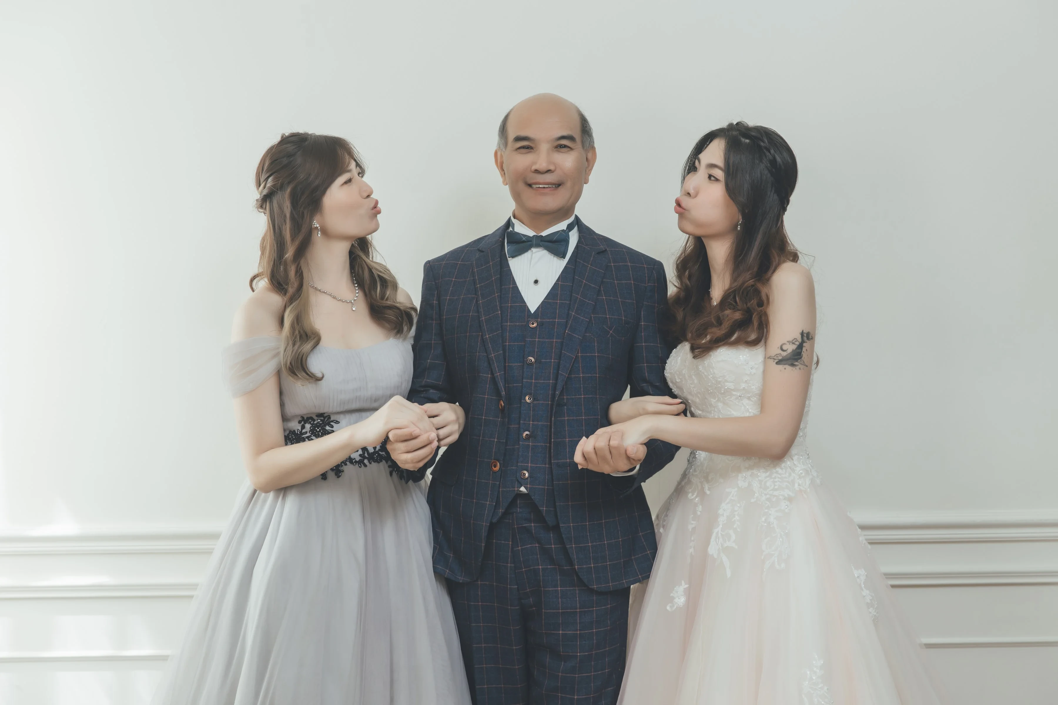 穿西裝的爸爸挽著兩個穿禮服的女兒，女兒們嘟嘴親他